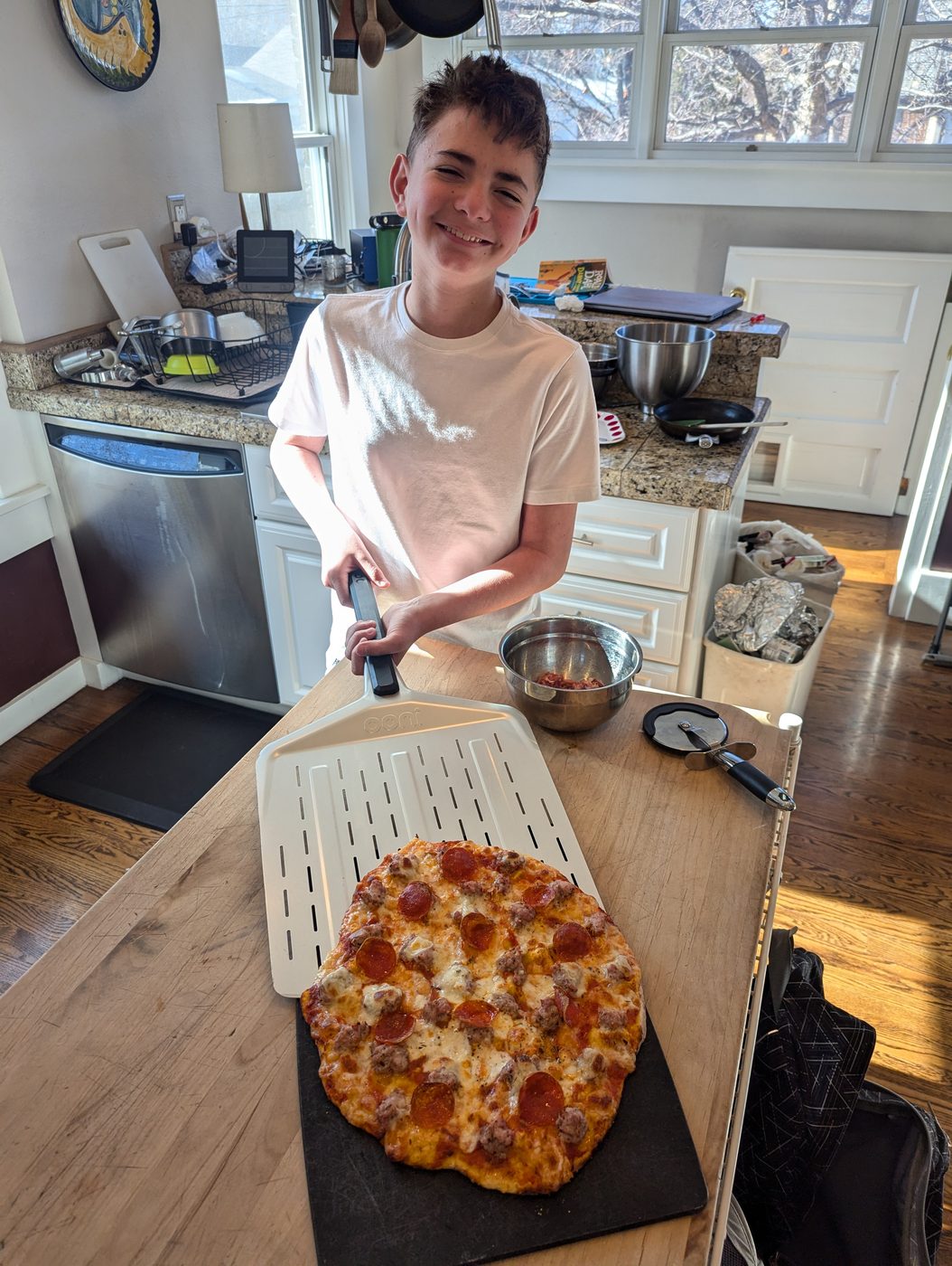 A young cook proudly holding a pizza peel behind a finished sausage-and-pepperoni pizza