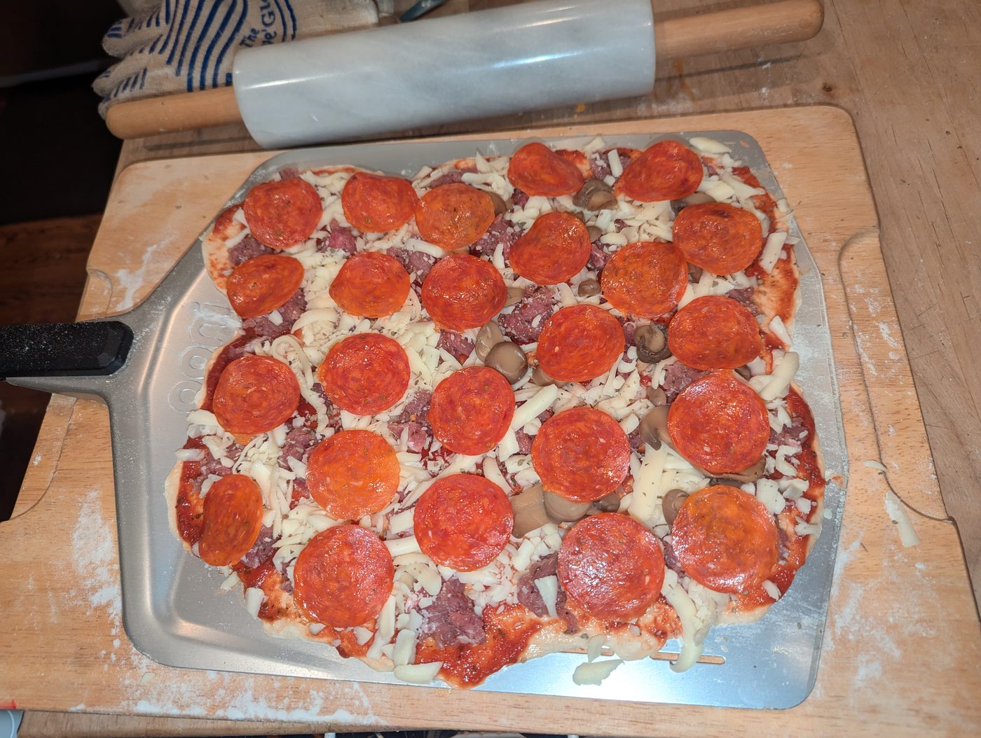 Fully topped pizza ready to bake: cheese, pepperoni, sausage, and mushrooms on a pizza peel