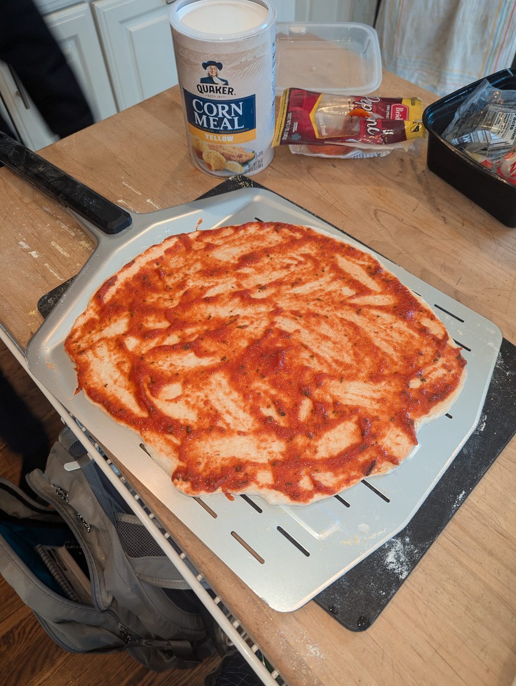 Pizza dough on a peel with sauce spread evenly all the way to the edge, cornmeal container in the background